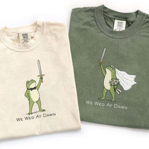 May include: Two t-shirts, one cream and one olive green, each featuring a graphic of a frog holding a sword and the text "WE WED AT DAWN". The cream shirt shows a frog in a bowtie, while the olive shirt shows a frog with a veil and flowers.