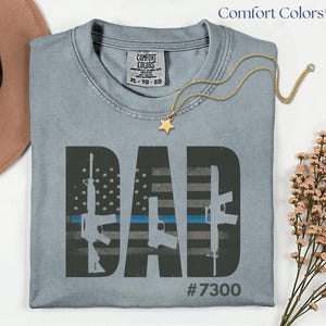 May include: A light blue Comfort Colors t-shirt with the word "DAD" in large, dark gray letters. The "A" in "DAD" is a stylized American flag with a blue line. The shirt also features three gun silhouettes and the number "7300". A gold necklace with a star pendant is draped on the shirt.
