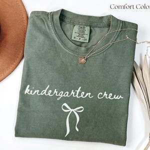 May include: An olive green t-shirt with the words "kindergarten crew" in white script and a white bow graphic. A gold necklace with a pendant is draped on the shirt. The brand name "Comfort Colors" is printed at the top right.