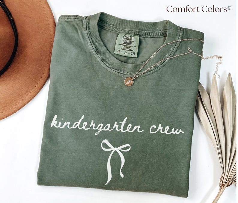 May include: An olive green t-shirt with the words "kindergarten crew" in white script and a white bow graphic. A gold necklace with a pendant is draped on the shirt. The brand name "Comfort Colors" is printed at the top right.