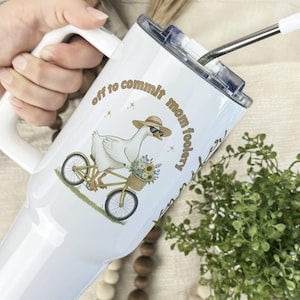 May include: White tumbler with a handle and a straw. The tumbler features a cartoon goose wearing sunglasses and a hat, riding a bicycle with a basket of flowers. Text on the tumbler reads "off to commit mom foolery."
