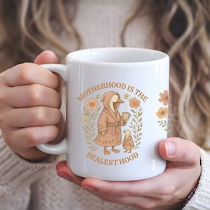 Puede incluir: Taza de cer&aacute;mica blanca con un dise&ntilde;o floral y un pato de dibujos animados que lleva una bata y sostiene una taza. El texto "Motherhood is the Realest Hood" est&aacute; impreso en la taza.