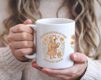 Peut inclure: Une tasse en céramique blanche avec un motif floral et un canard de dessin animé portant une robe de chambre et tenant une tasse. Le texte "Motherhood is the Realest Hood" est imprimé sur la tasse.