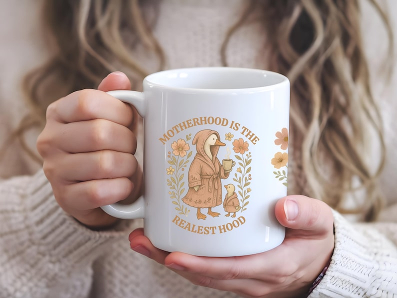 Puede incluir: Taza de cer&aacute;mica blanca con un dise&ntilde;o floral y un pato de dibujos animados que lleva una bata y sostiene una taza. El texto "Motherhood is the Realest Hood" est&aacute; impreso en la taza.