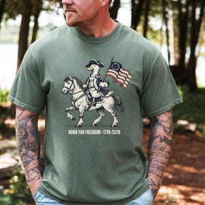 May include: Olive green t-shirt featuring a cartoon duck riding a horse and holding a flag. The duck is dressed in colonial attire. The flag has stars and stripes. The text "HONK FOR FREEDOM - 1776-2026" is below the image.