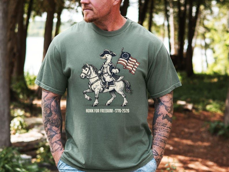 May include: Olive green t-shirt featuring a cartoon duck riding a horse and holding a flag. The duck is dressed in colonial attire. The flag has stars and stripes. The text "HONK FOR FREEDOM - 1776-2026" is below the image.