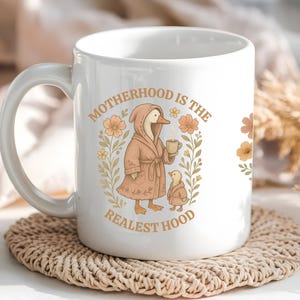 Puede incluir: Taza de cer&aacute;mica blanca con un pato rosa con una bata y sosteniendo una taza de caf&eacute;. La taza tiene el texto "Motherhood is the Realest Hood" con ilustraciones florales.