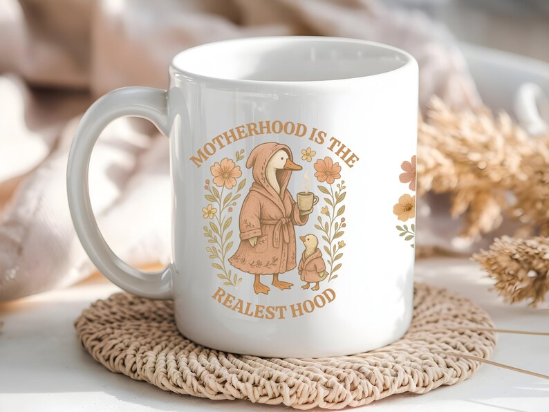 Puede incluir: Taza de cer&aacute;mica blanca con un pato rosa con una bata y sosteniendo una taza de caf&eacute;. La taza tiene el texto "Motherhood is the Realest Hood" con ilustraciones florales.