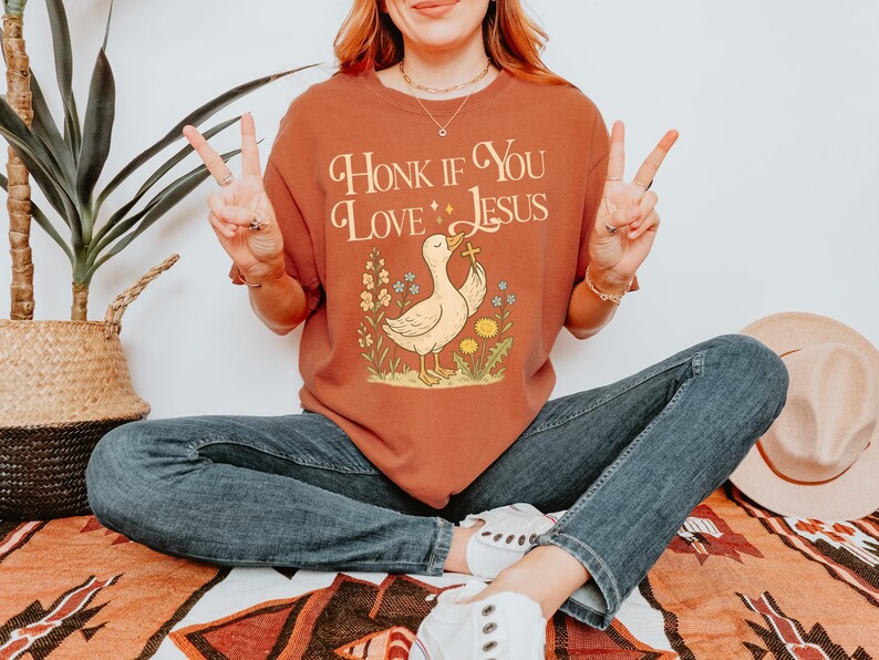 Puede incluir: Camiseta de color &oacute;xido con la frase "Honk If You Love Jesus" sobre un pato de dibujos animados que sostiene una cruz. El pato est&aacute; rodeado de flores. La persona lleva vaqueros azules y zapatillas blancas.