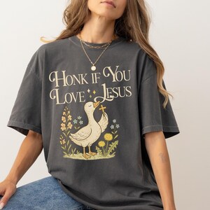Puede incluir: Camiseta gris oscuro con el texto "Honk If You Love Jesus" y un dibujo de un pato sosteniendo una cruz, rodeado de flores. La camiseta tiene mangas cortas y un corte holgado.