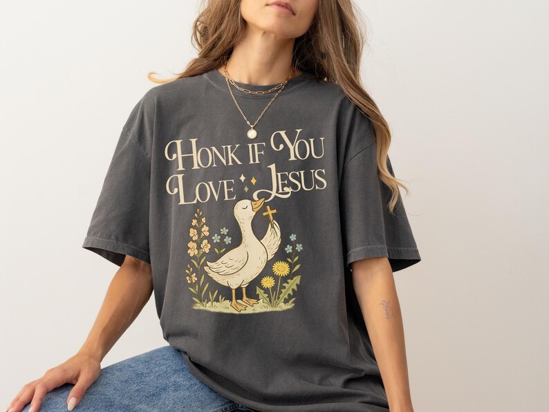 Puede incluir: Camiseta gris oscuro con el texto "Honk If You Love Jesus" y un dibujo de un pato sosteniendo una cruz, rodeado de flores. La camiseta tiene mangas cortas y un corte holgado.