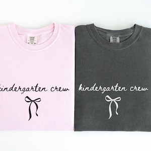 May include: Two folded t-shirts, one light pink and one charcoal gray, are displayed. Each shirt features the words "kindergarten crew" in a cursive font, with a bow graphic below. The shirts are likely for a school or group.