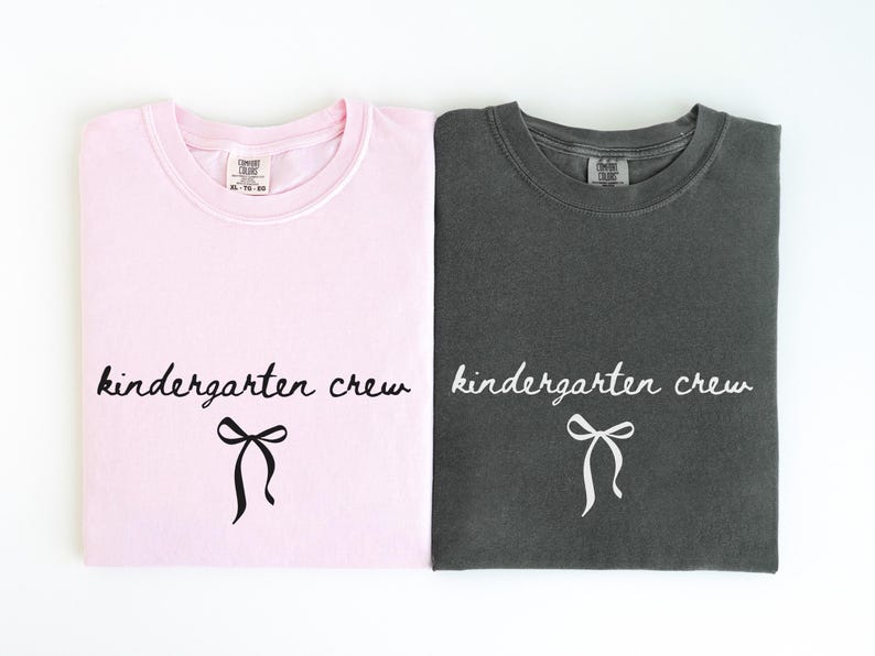 May include: Two folded t-shirts, one light pink and one charcoal gray, are displayed. Each shirt features the words "kindergarten crew" in a cursive font, with a bow graphic below. The shirts are likely for a school or group.