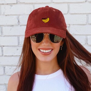 Puede incluir: Una mujer con una gorra de b&eacute;isbol de pana marr&oacute;n con el texto "That's" y un dise&ntilde;o de pl&aacute;tano bordado en amarillo. Tambi&eacute;n lleva gafas de sol y un top blanco sin mangas.