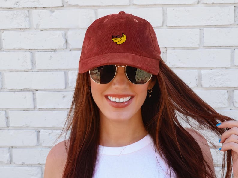 Puede incluir: Una mujer con una gorra de b&eacute;isbol de pana marr&oacute;n con el texto "That's" y un dise&ntilde;o de pl&aacute;tano bordado en amarillo. Tambi&eacute;n lleva gafas de sol y un top blanco sin mangas.