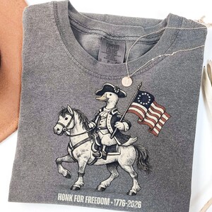 May include: Gray t-shirt featuring a cartoon duck riding a horse and holding a flag. The duck is dressed in a colonial outfit. The flag has 13 stars and stripes. The text "HONK FOR FREEDOM - 1776-2026" is printed at the bottom.