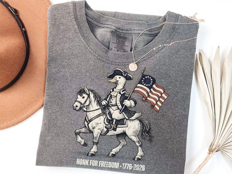 May include: Gray t-shirt featuring a cartoon duck riding a horse and holding a flag. The duck is dressed in a colonial outfit. The flag has 13 stars and stripes. The text "HONK FOR FREEDOM - 1776-2026" is printed at the bottom.