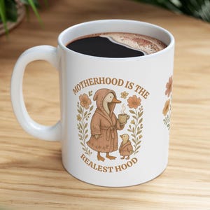 Puede incluir: Taza de caf&eacute; de cer&aacute;mica blanca con un pato marr&oacute;n que lleva una bata y sostiene una taza de caf&eacute;. La taza tiene el texto "Motherhood is the Realest Hood" con ilustraciones florales.