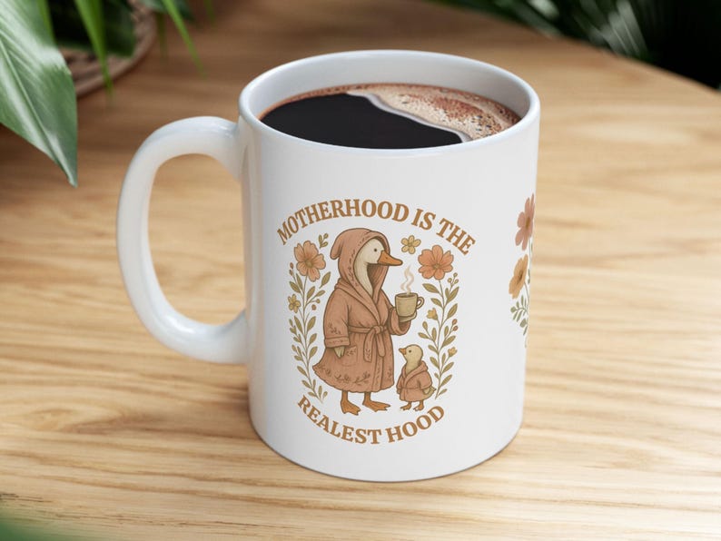 Puede incluir: Taza de caf&eacute; de cer&aacute;mica blanca con un pato marr&oacute;n que lleva una bata y sostiene una taza de caf&eacute;. La taza tiene el texto "Motherhood is the Realest Hood" con ilustraciones florales.