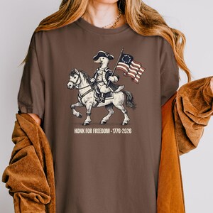 May include: Brown t-shirt featuring a cartoon duck riding a horse and holding a flag. The duck is dressed in colonial attire. The flag has a circle of stars. The text "HONK FOR FREEDOM - 1776-2026" is below the image.