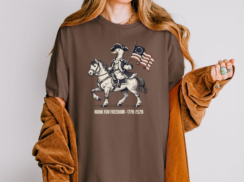 May include: Brown t-shirt featuring a cartoon duck riding a horse and holding a flag. The duck is dressed in colonial attire. The flag has a circle of stars. The text "HONK FOR FREEDOM - 1776-2026" is below the image.