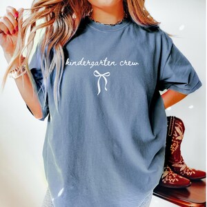 May include: A dusty blue Comfort Colors t-shirt with the words "kindergarten crew" in white script, along with a white bow graphic. The shirt is a relaxed fit.