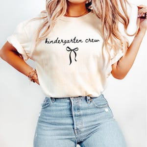 May include: Cream-colored t-shirt with the words "kindergarten crew" in black script, accompanied by a black bow graphic. The shirt is paired with light blue denim jeans.