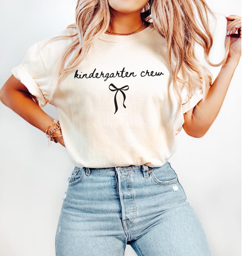 May include: Cream-colored t-shirt with the words "kindergarten crew" in black script, accompanied by a black bow graphic. The shirt is paired with light blue denim jeans.