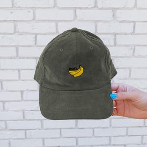 Puede incluir: Una gorra de b&eacute;isbol de pana verde con un dise&ntilde;o bordado de pl&aacute;tanos amarillos y el texto "that's" en la parte delantera.