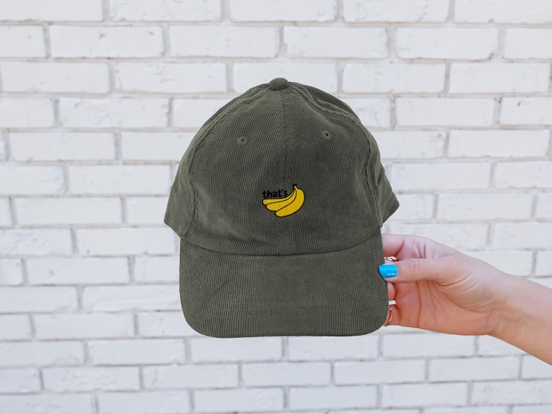 Puede incluir: Una gorra de b&eacute;isbol de pana verde con un dise&ntilde;o bordado de pl&aacute;tanos amarillos y el texto "that's" en la parte delantera.
