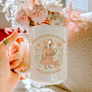 Puede incluir: Taza de cer&aacute;mica blanca con fondo azul claro y dise&ntilde;o floral. La taza presenta un ganso de dibujos animados con una bata rosa y sosteniendo una taza de caf&eacute;. El texto "Motherhood is the Realest Hood" est&aacute; impreso en la taza.