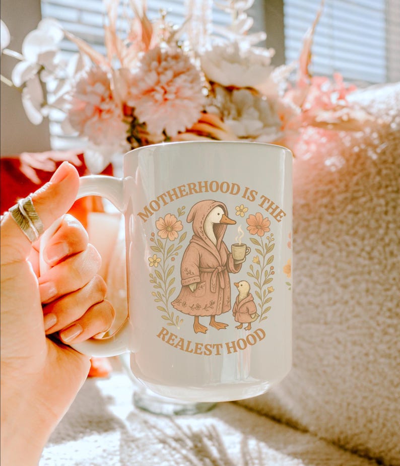 Puede incluir: Taza de cer&aacute;mica blanca con fondo azul claro y dise&ntilde;o floral. La taza presenta un ganso de dibujos animados con una bata rosa y sosteniendo una taza de caf&eacute;. El texto "Motherhood is the Realest Hood" est&aacute; impreso en la taza.