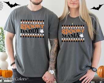 May include: Two gray Comfort Colors t-shirts with Halloween-themed text. One shirt says "Spooky Daddy" and the other says "Spooky Mama", both with a ghost graphic. The shirts have a black, white, and orange checkered design.