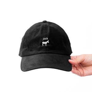 May include: Black corduroy baseball cap with a curved brim. The cap features the word "jack" above a white donkey graphic. The hat is held by a person's hand against a white background.