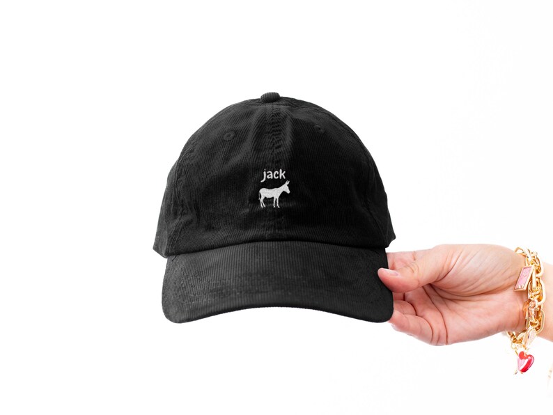 May include: Black corduroy baseball cap with a curved brim. The cap features the word "jack" above a white donkey graphic. The hat is held by a person's hand against a white background.