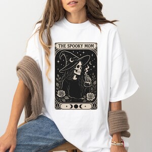 May include: White t-shirt featuring a black and cream graphic of a skeleton witch holding a potion bottle, with the text "THE SPOOKY MOM". The design includes a witch hat, roses, and crescent moons.