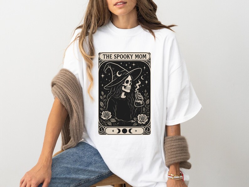 May include: White t-shirt featuring a black and cream graphic of a skeleton witch holding a potion bottle, with the text "THE SPOOKY MOM". The design includes a witch hat, roses, and crescent moons.