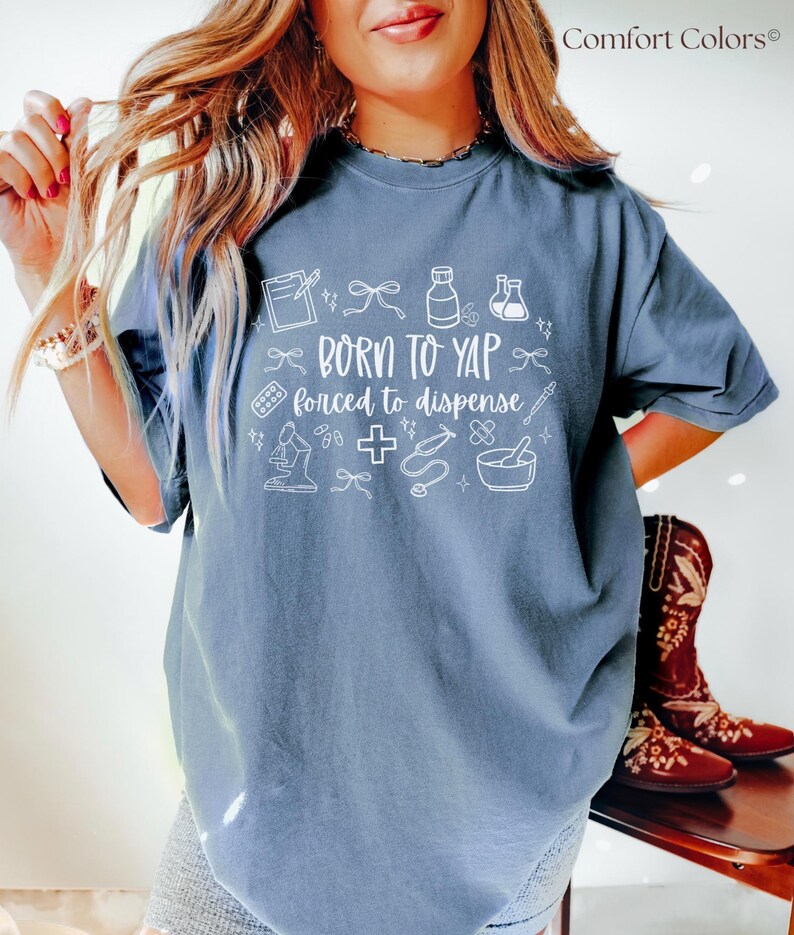 May include: A Comfort Colors blue-grey t-shirt with white graphic text and medical-themed illustrations. The text reads "Born to Yap, forced to dispense." The shirt features a crew neck and short sleeves.
