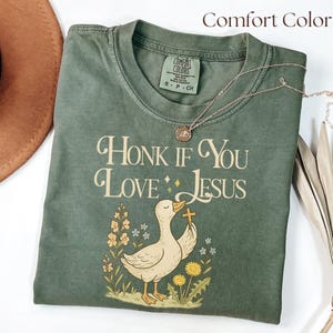 A folded sage green Comfort Colors T-shirt is displayed on a white surface beside a brown felt hat and neutral-toned feathers. The shirt features a whimsical illustration of a goose holding a cross in its beak, surrounded by yellow and peach flowers. Above the design, vintage-style text reads “Honk If You Love Jesus” in cream and gold lettering. A dainty necklace rests on top.