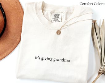 May include: Cream-colored t-shirt with the phrase "it's giving grandma" in black lettering. A gold necklace with a circular pendant rests on the shirt. The shirt is from Comfort Colors.