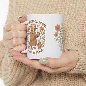 Puede incluir: Taza de cer&aacute;mica blanca con un pato y un patito marrones que llevan batas y sostienen tazas. El texto dice "Motherhood is the Realest Hood" con ilustraciones florales.