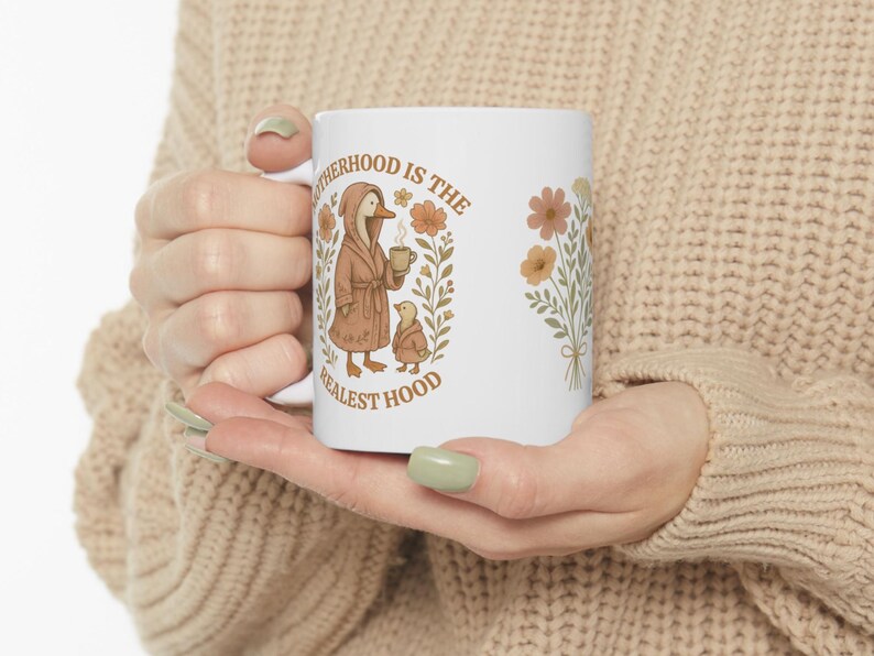 Puede incluir: Taza de cer&aacute;mica blanca con un pato y un patito marrones que llevan batas y sostienen tazas. El texto dice "Motherhood is the Realest Hood" con ilustraciones florales.
