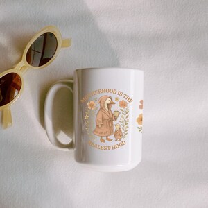Puede incluir: Taza de cer&aacute;mica blanca con un pato marr&oacute;n que lleva una bata y sostiene una taza. El texto en la taza dice "Motherhood is the Realest Hood" con detalles florales.