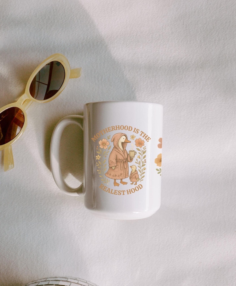 Puede incluir: Taza de cer&aacute;mica blanca con un pato marr&oacute;n que lleva una bata y sostiene una taza. El texto en la taza dice "Motherhood is the Realest Hood" con detalles florales.