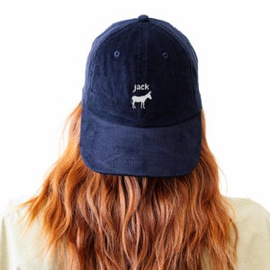 May include: Navy blue corduroy baseball cap with the word "jack" and a white donkey graphic embroidered on the front. The cap has a curved brim and a button on top. The hat is worn by a person with long, wavy red hair.