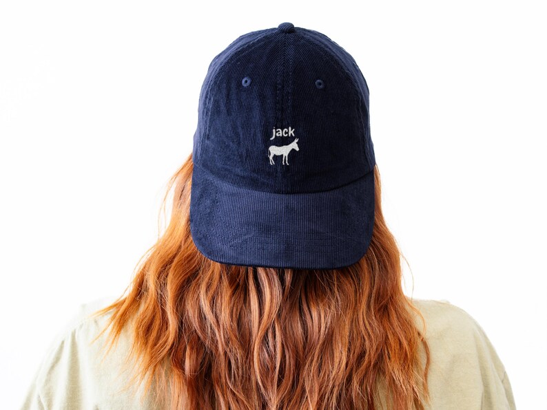 May include: Navy blue corduroy baseball cap with the word "jack" and a white donkey graphic embroidered on the front. The cap has a curved brim and a button on top. The hat is worn by a person with long, wavy red hair.