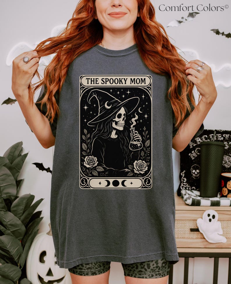 Woman wearing a dark gray oversized t-shirt featuring a gothic-style Halloween design labeled “The Spooky Mom,” with a skeleton witch holding a bubbling potion. The shirt includes celestial elements, roses, and moon phases in a tarot card-style frame. Surrounding the model is festive Halloween decor including a ghost plush, skull, and bats.