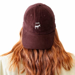 May include: A corduroy baseball cap in a deep burgundy color. The cap features the word "jack" embroidered above a white donkey silhouette. The cap is worn by a person with long, auburn hair.
