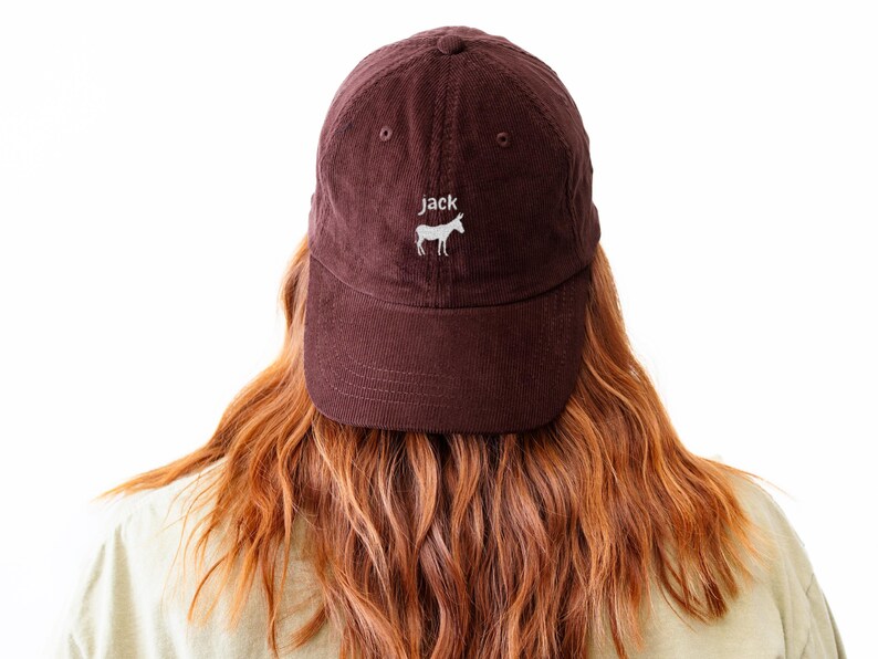 May include: A corduroy baseball cap in a deep burgundy color. The cap features the word "jack" embroidered above a white donkey silhouette. The cap is worn by a person with long, auburn hair.