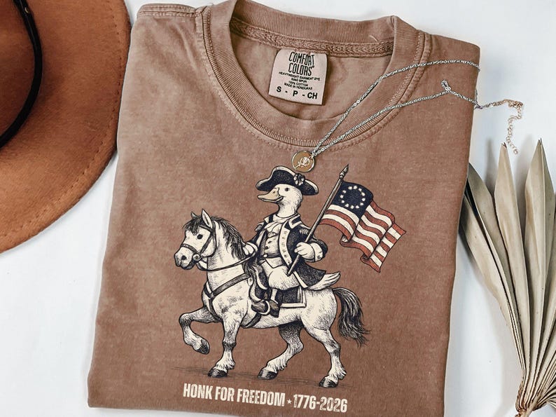 May include: Brown Comfort Colors t-shirt featuring a duck dressed as a revolutionary war figure riding a horse and holding a flag. The shirt has the text "HONK FOR FREEDOM - 1776-2026" at the bottom. A silver necklace and a hat are also visible.